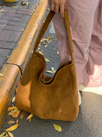 Brown suede handbag held by a person on a sidewalk with autumn leaves.