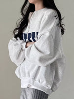 A woman wearing an oversized light gray heather sweatshirt with a navy blue and white graphic across the chest, paired with gray ribbed pants, set against a plain light gray background.