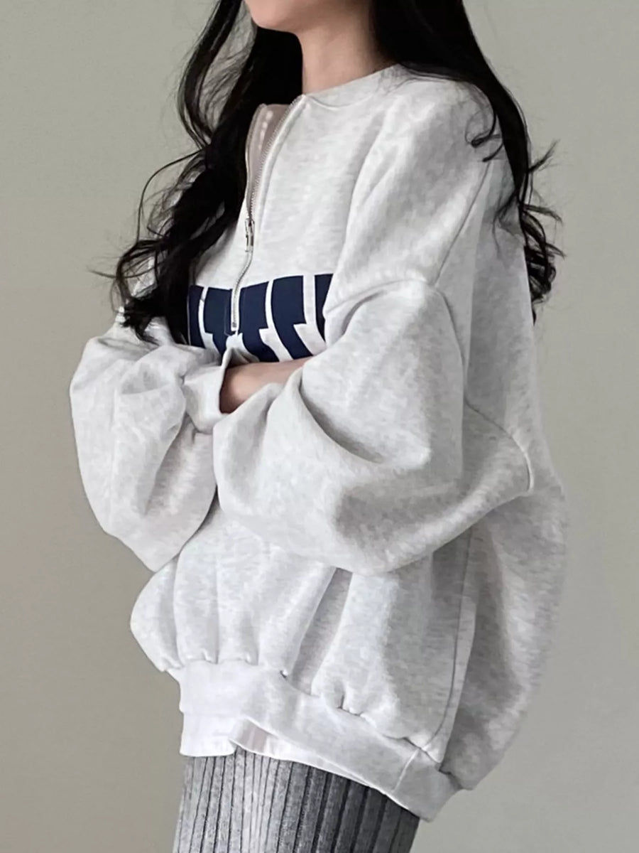 A woman wearing an oversized light gray heather sweatshirt with a navy blue and white graphic across the chest, paired with gray ribbed pants, set against a plain light gray background.