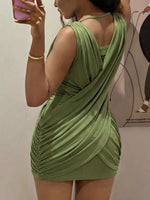 A woman wearing a ruched sage green mini dress, photographed in a mirror selfie. The dress features draped, gathered fabric details and a crossover wrap-style back.