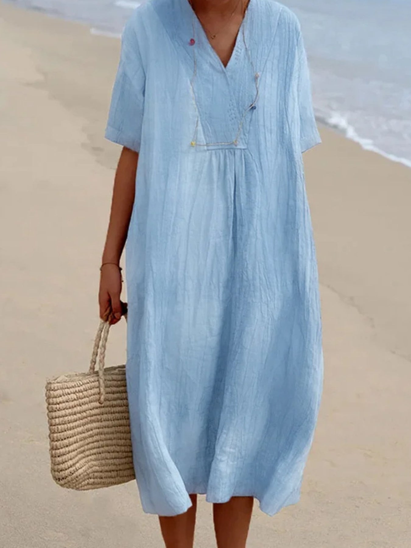 Person wearing a light blue dress on a beach with a straw bag.