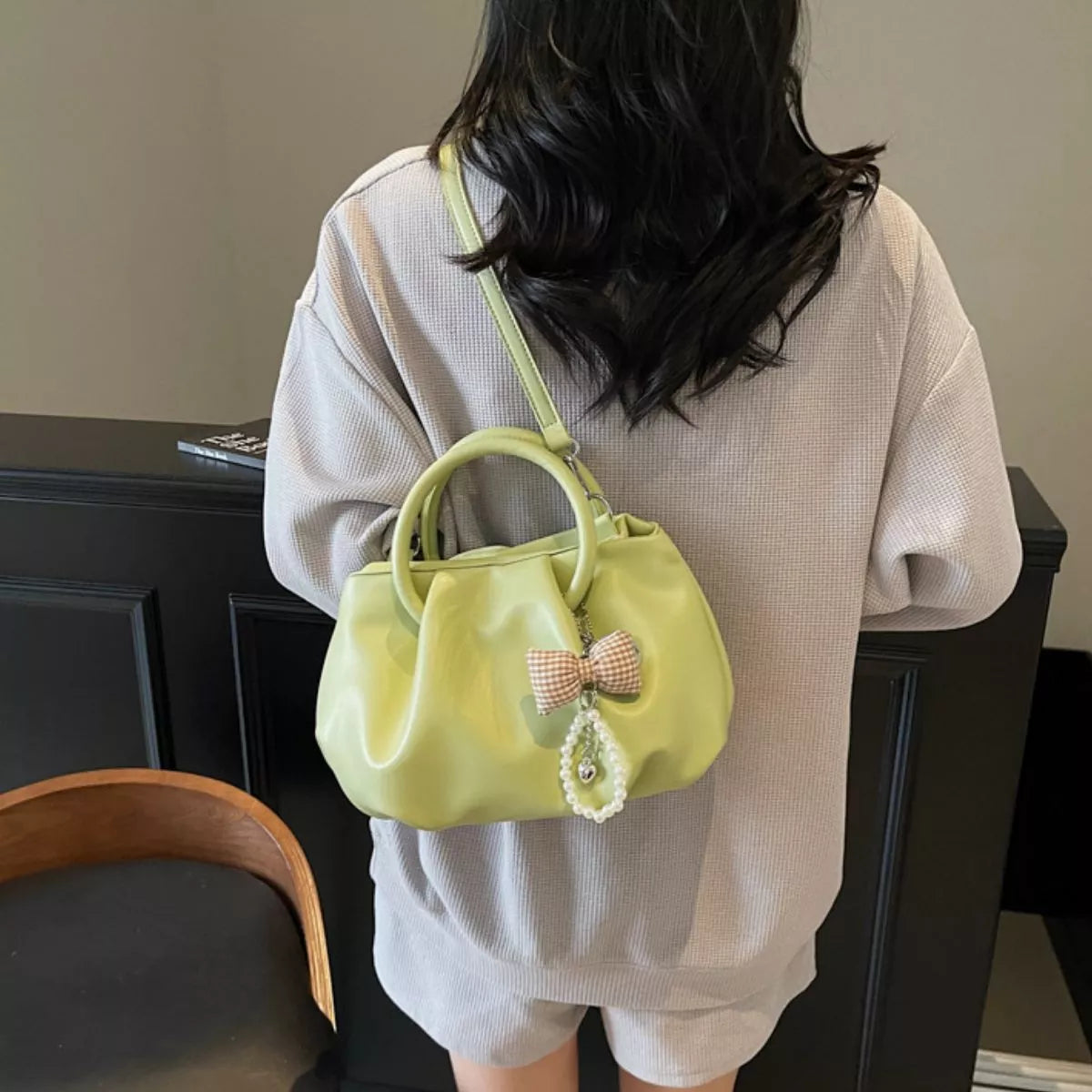A woman from behind wearing a light beige knit sweater and shorts, carrying a small light green leather handbag with a top handle, a pink-and-white checkered bow, and a pearl charm.