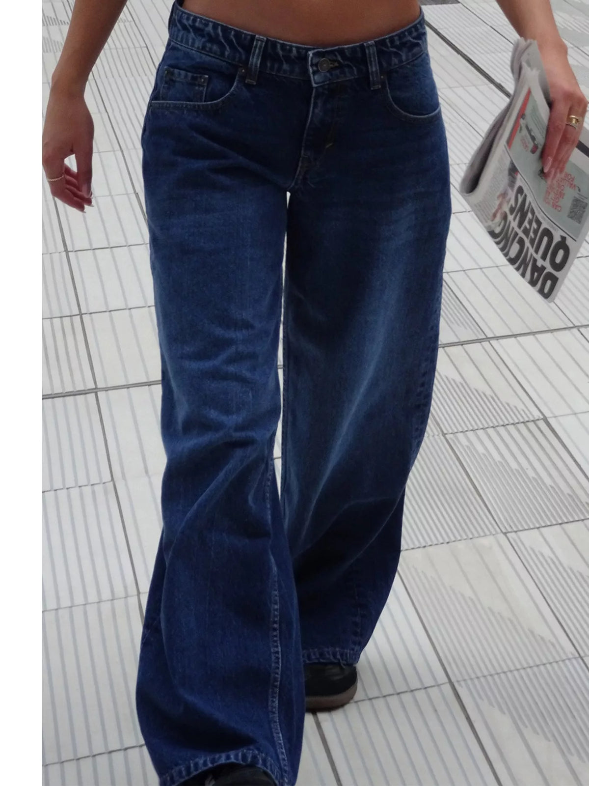 A person wearing dark blue wide-leg denim jeans, shown from the waist down while walking on a light gray tiled floor and holding a gray newspaper.