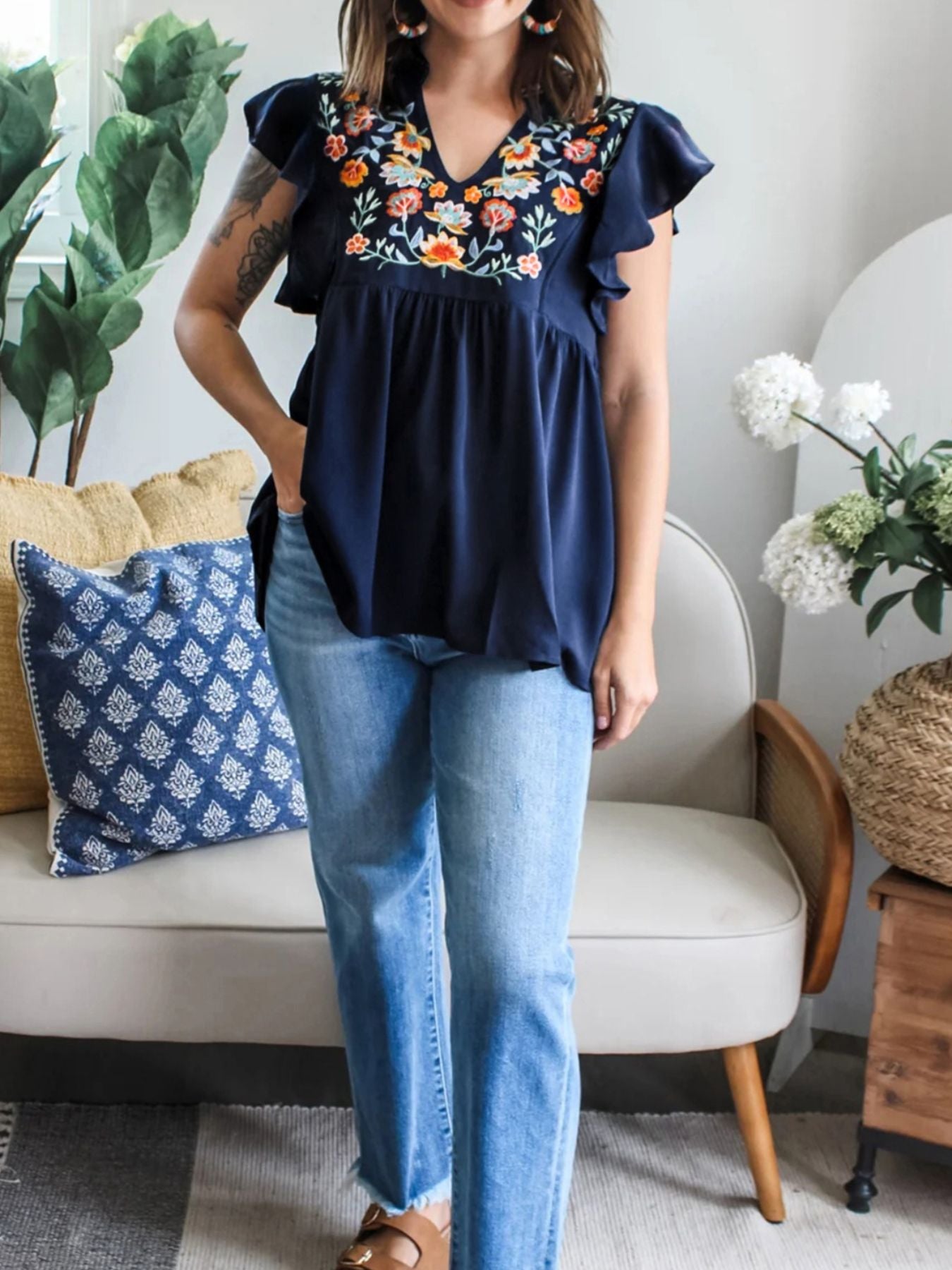 Woman wearing a navy blue top with floral embroidery and blue jeans in a living room setting.