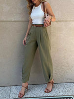 A woman models a pair of high-waisted olive green wide-leg trousers with a matching brown belt, paired with a white tank top and brown strappy heels, standing against a beige stone wall.