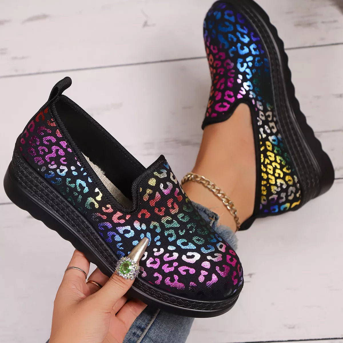 Black slip-on shoes with colorful leopard print pattern held by a hand on a light wooden floor.