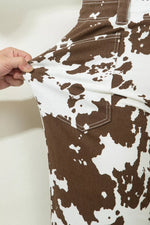 A close-up of a brown and white cow-print garment being held by a hand to show the fabric texture and stretch, featuring a chest pocket with white contrast stitching.