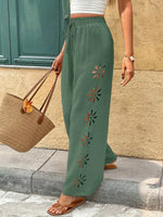 Person wearing green pants with floral embroidery, standing against a stone wall.
