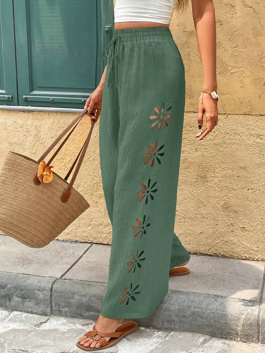 Person wearing green pants with floral embroidery, standing against a stone wall.