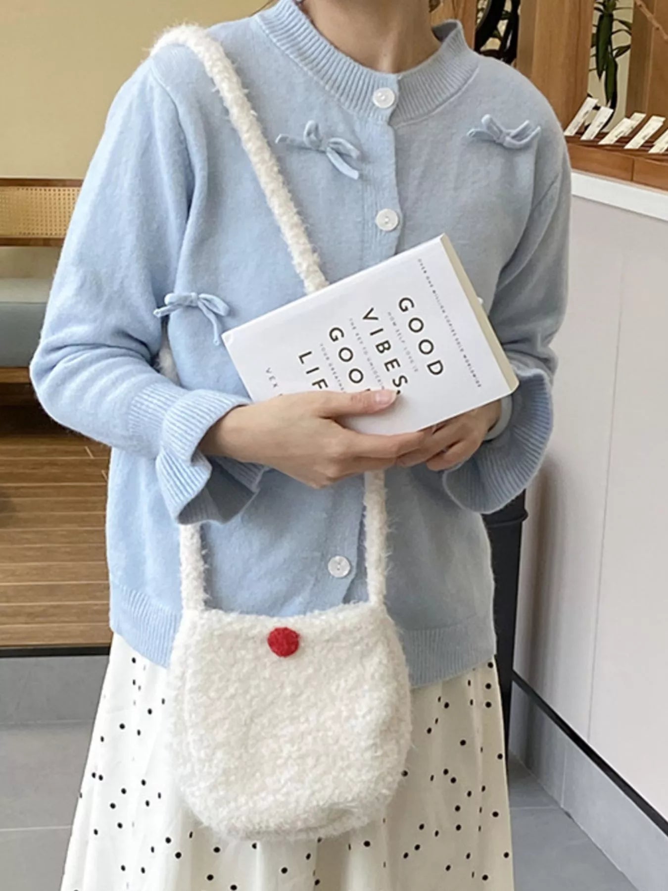 Person holding a white textured bag with a red button and a book titled 'Good Vibes Good Life'.