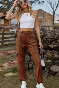 Woman in white top and brown cargo pants standing outdoors near a stone wall.
