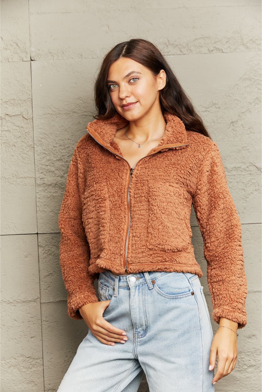 Woman wearing a brown fuzzy jacket and light blue jeans against a neutral wall.