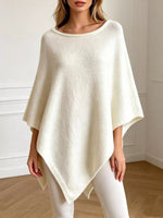 A woman wearing a cream-colored, open-knit poncho sweater with a scoop neckline and draped sleeves, paired with white leggings, standing in a brightly lit room with white walls and a light wood floor.