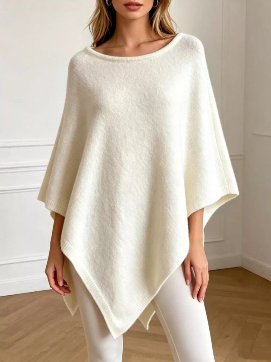 A woman wearing a cream-colored, open-knit poncho sweater with a scoop neckline and draped sleeves, paired with white leggings, standing in a brightly lit room with white walls and a light wood floor.