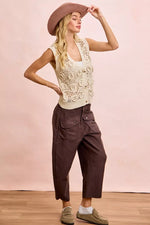 Woman wearing a lace top, wide-brimmed hat, and brown pants against a pink background