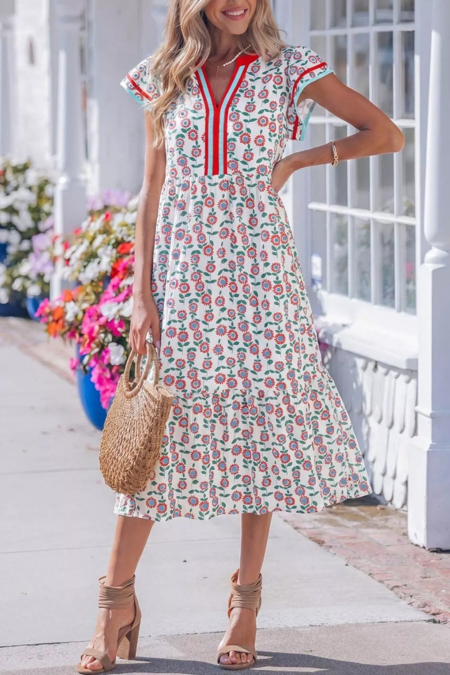 Woman wearing a floral dress with red accents, standing on a sidewalk with flowers in the background.