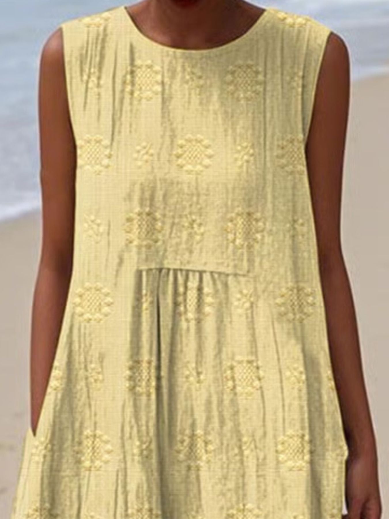 Light yellow sleeveless dress with intricate patterns worn by a person on a beach.