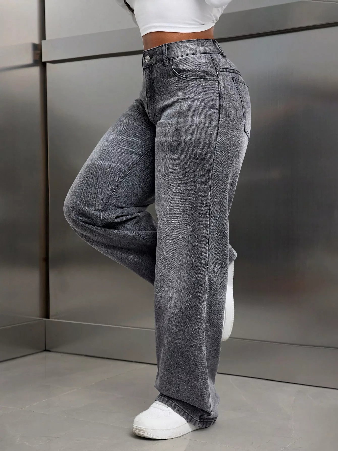 Person wearing gray high-waisted jeans and white sneakers against a metallic wall.