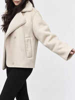 A woman wearing a cropped beige shearling-style jacket with a soft collar and front buttons, paired with black pants, against a plain light gray studio background.