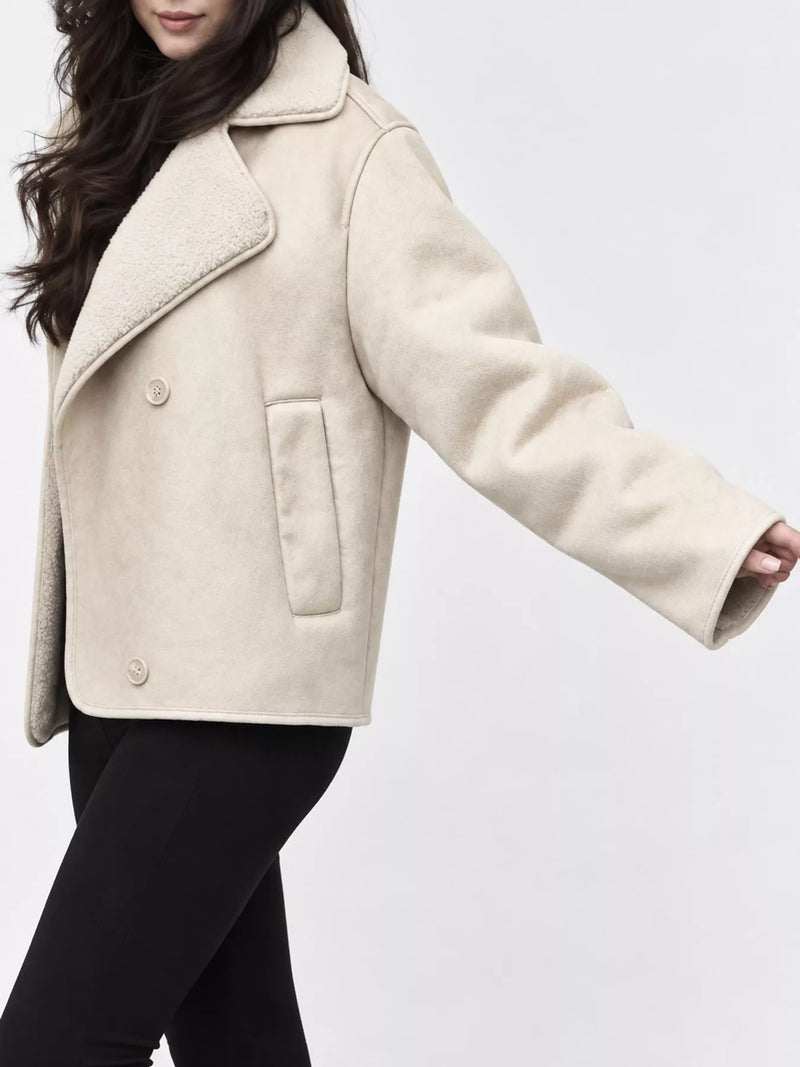 A woman wearing a cropped beige shearling-style jacket with a soft collar and front buttons, paired with black pants, against a plain light gray studio background.