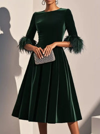 Green velvet dress with feathered sleeves held by a hand with a clutch against a neutral background