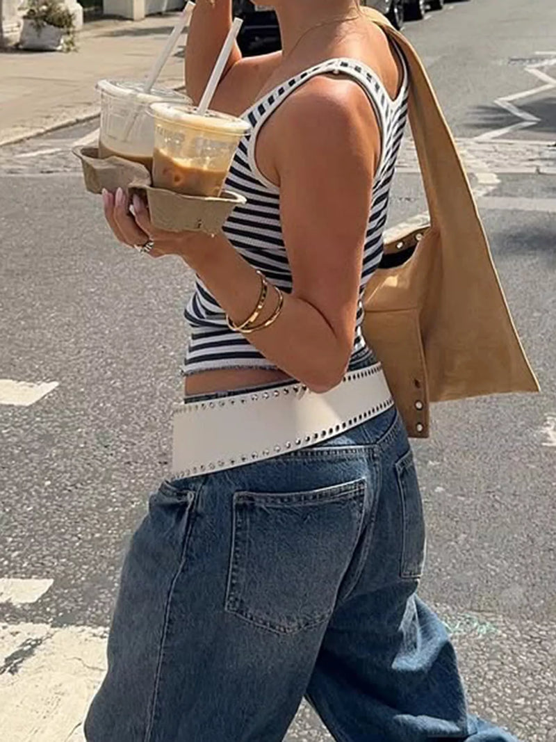 Person wearing a striped tank top and jeans, holding a drink on a street.