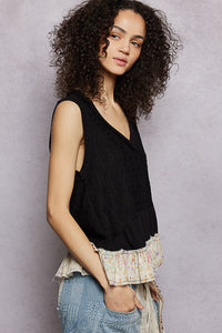 Woman wearing a black sleeveless top with floral trim and blue jeans against a gray background