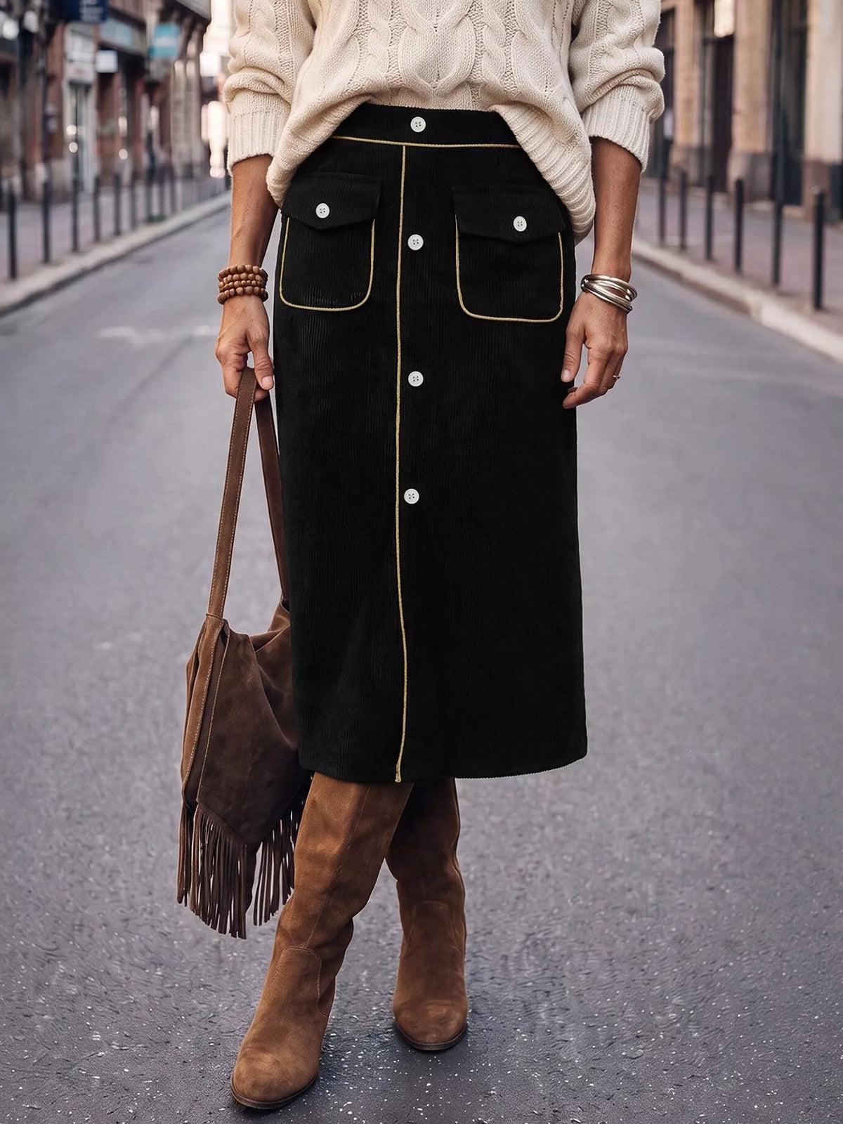 A person standing on a city street wearing a black midi skirt with gold contrast piping and front buttons, a cream cable-knit sweater, and brown suede knee-high boots, while carrying a brown fringe shoulder bag.