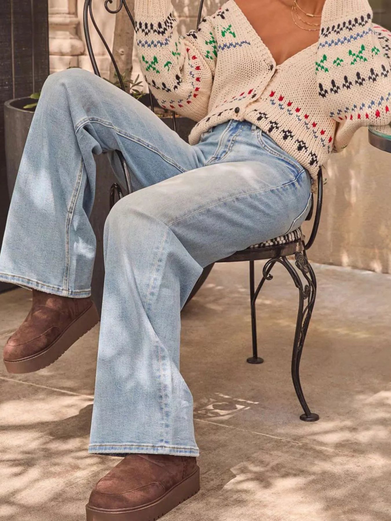 Person wearing a patterned cardigan, light blue jeans, and brown shoes sitting on a chair.