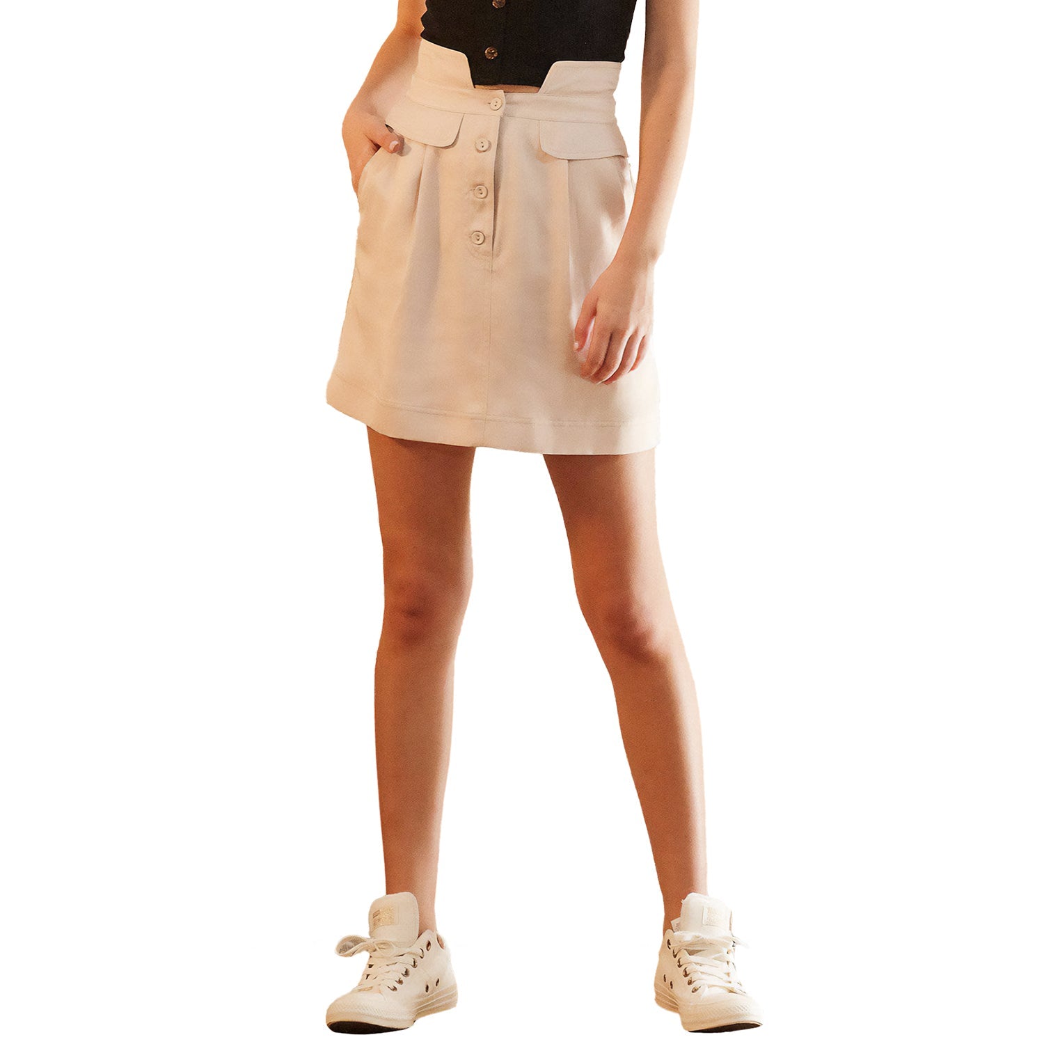 A woman wearing a high waisted beige utility skirt with side pockets and a button fly. She is also wearing a black top and white sneakers.