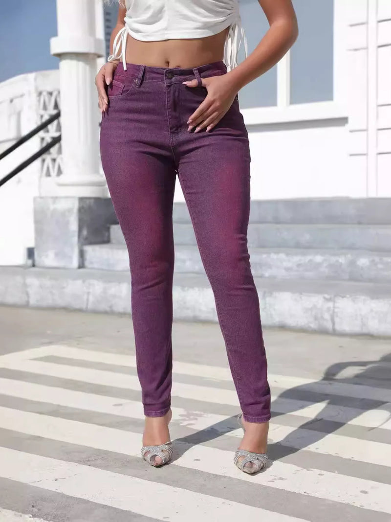 A model wearing high-waisted, ankle-length purple skinny jeans with a faded wash, styled with a white crop top and patterned heels, standing on outdoor concrete steps.