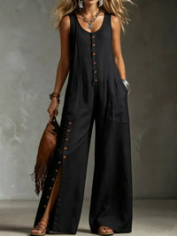 Woman wearing a black sleeveless jumpsuit with buttons, holding a brown leather bag against a gray background.