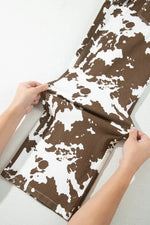 A pair of shorts with a brown and white cow print pattern, being held open by a person's hands against a light gray background.
