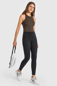 Woman wearing a brown tank top and black leggings holding a tennis racket on a white background