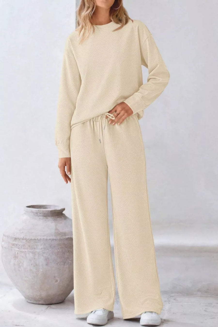 Person wearing a beige outfit standing next to a textured vase on a light background