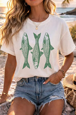Person wearing a white t-shirt with green fish graphics on a beach.