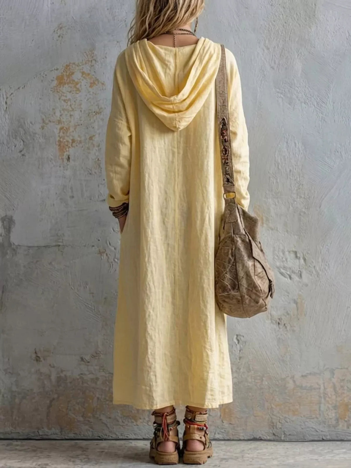 A woman viewed from behind wearing a pale yellow hooded maxi dress with long sleeves and pockets, carrying a brown shoulder bag and wearing brown lace-up sandals, standing against a textured gray wall.