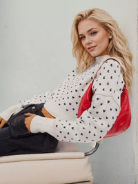 Woman sitting on a chair wearing a white sweater with black patterns and holding a red handbag.