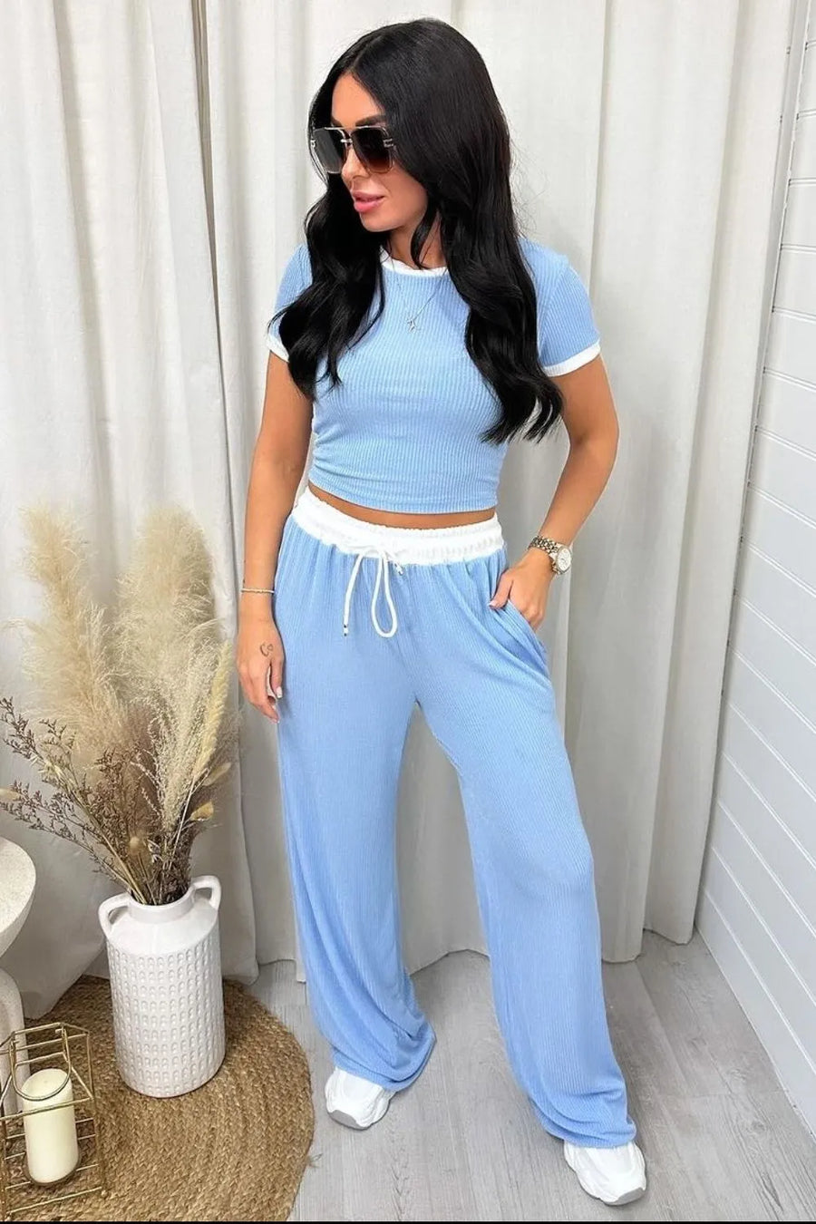 Woman wearing a blue outfit with white pants, standing indoors against a neutral background.