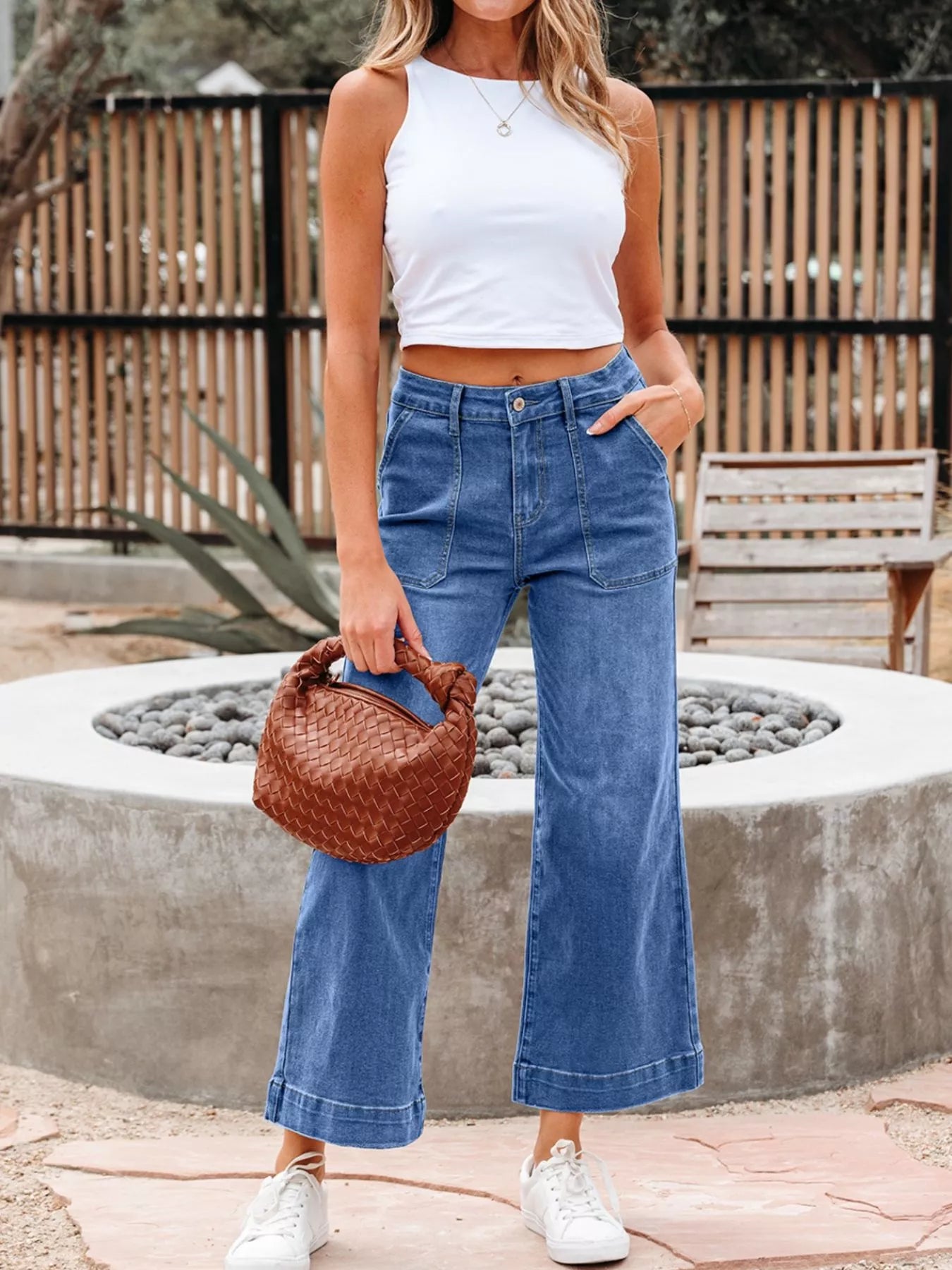 Woman wearing a white tank top and blue jeans holding a brown woven handbag outdoors.