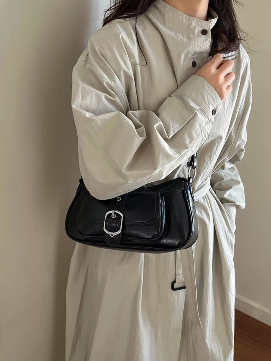 A person wearing a light beige oversized shirt dress carries a black leather shoulder bag with a silver buckle, standing against a plain beige wall.