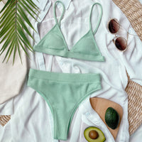Green bikini set on a white surface with sunglasses, avocado, and palm leaves.