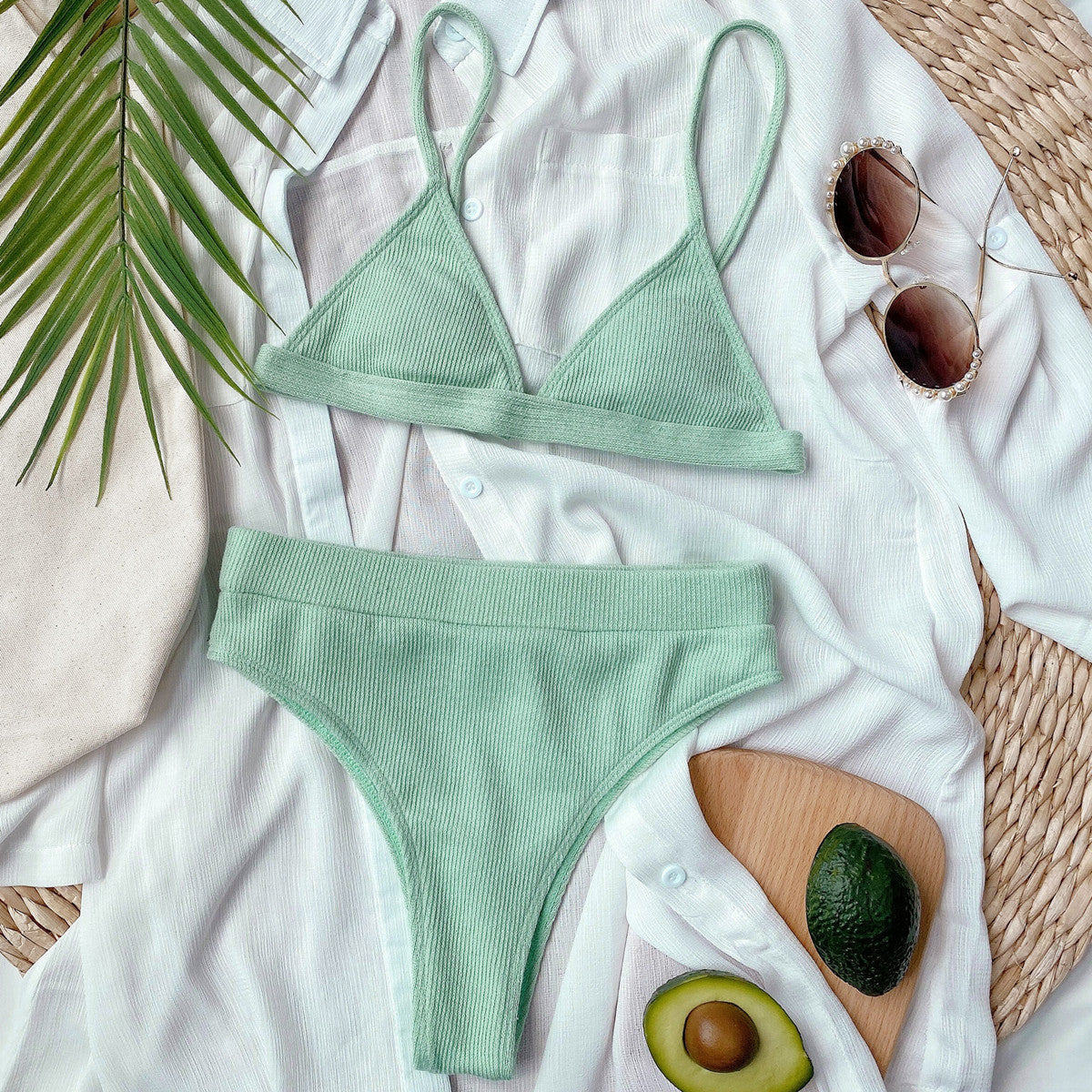 Green bikini set on a white surface with sunglasses, avocado, and palm leaves.