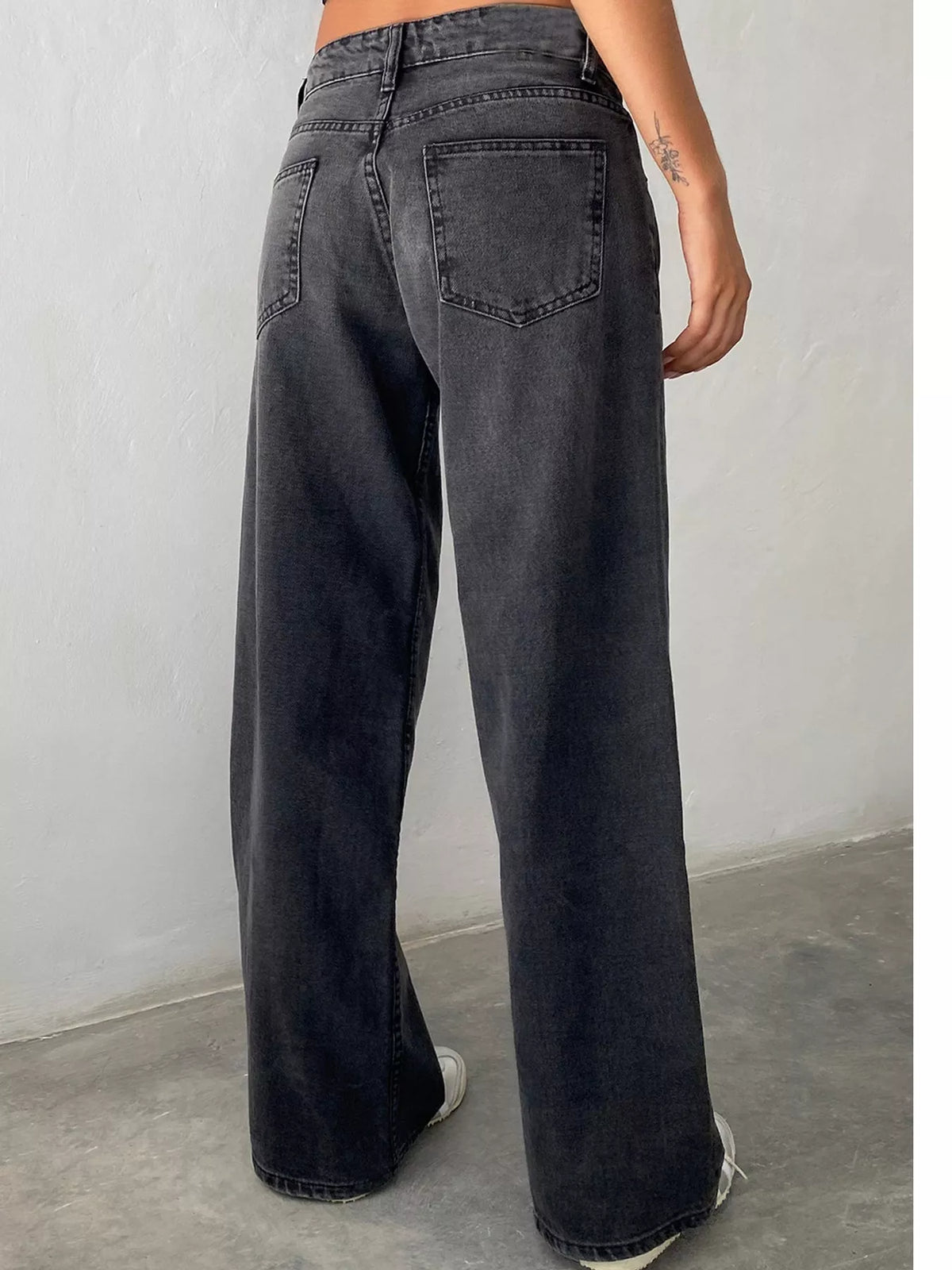 A person shown from the back wearing dark gray wide-leg jeans, standing against a light gray wall and concrete floor.