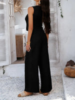 Woman wearing a black sleeveless top and wide-leg pants standing in a sunlit room with white walls and wooden furniture.