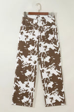 A pair of wide-leg jeans with a brown and white abstract splatter pattern, hanging on a wooden hanger against a plain off-white wall.