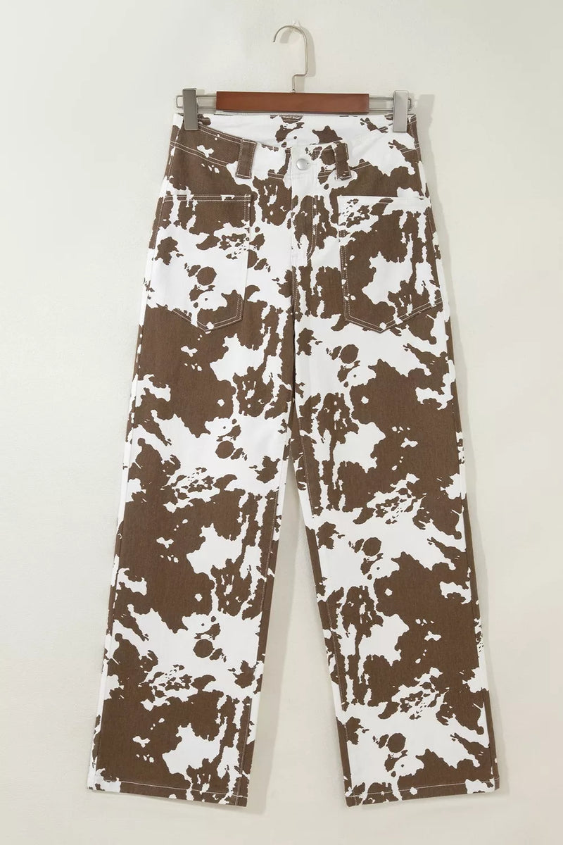 A pair of wide-leg jeans with a brown and white abstract splatter pattern, hanging on a wooden hanger against a plain off-white wall.