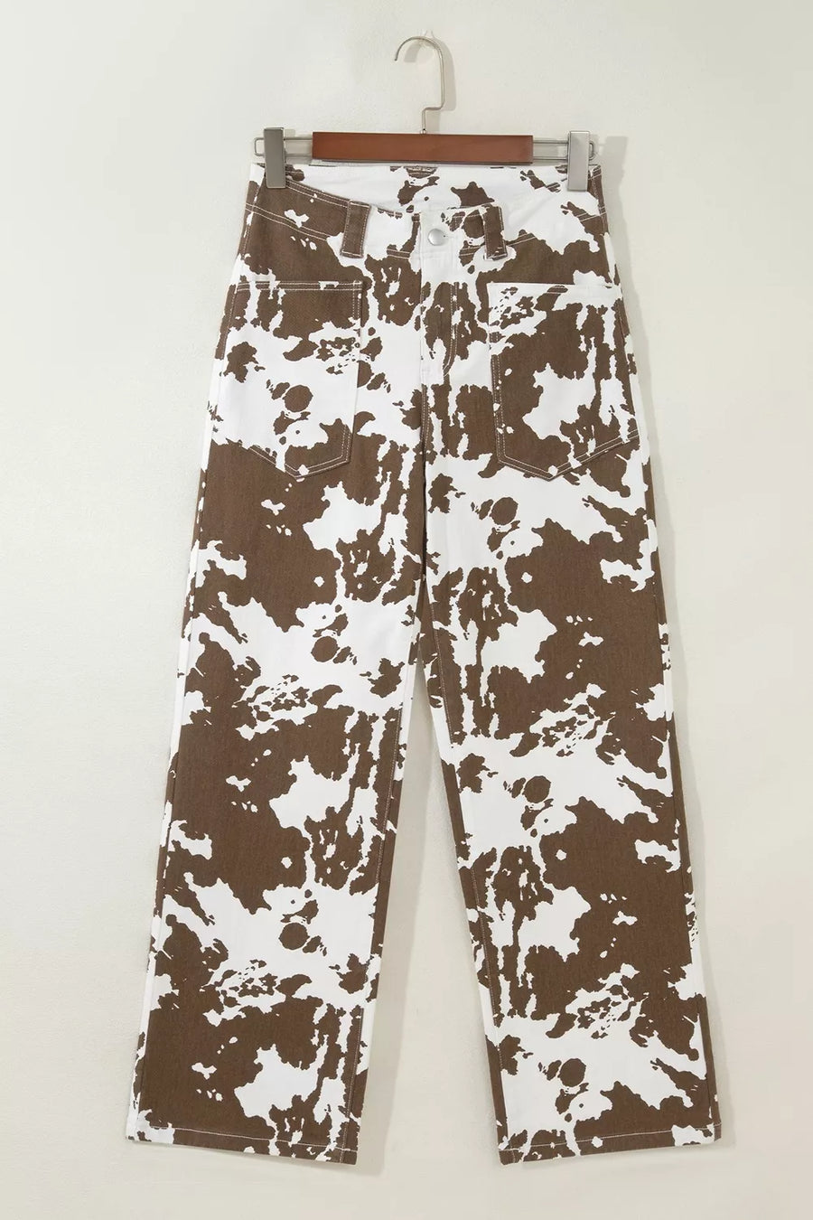 A pair of wide-leg jeans with a brown and white abstract splatter pattern, hanging on a wooden hanger against a plain off-white wall.
