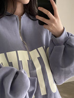 A close-up of a person wearing a lavender-gray quarter-zip sweatshirt with large cream lettering on the front, holding a black smartphone in a mirror selfie.