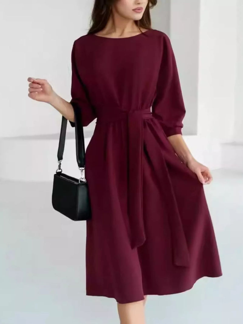 A woman wearing a knee-length burgundy dress with elbow-length sleeves, a boat neckline, and a self-tie waist belt. She is holding a small black shoulder bag against a clean, light gray studio background.